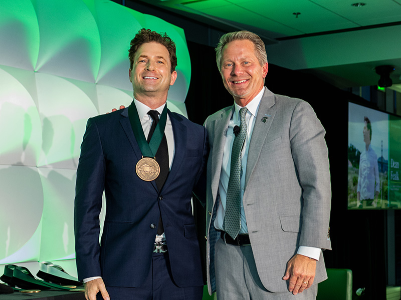 Dean Fealk and President Kevin Guskiewicz in suits stand side by side on a stage with green and white lighting.