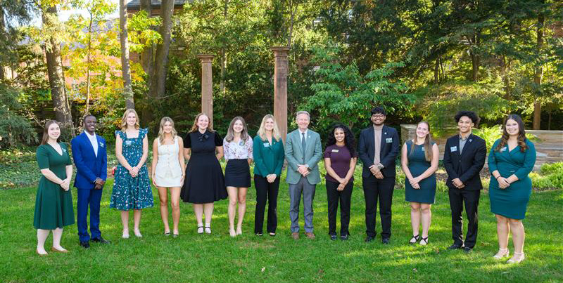 The 2025 MSU Homecoming Court poses for a group photo outdoors on campus, standing in a row on a sunny day with trees and brick columns in the background. Members wear a mix of formal and semi-formal attire in MSU colors, joined at the center by MSU President Kevin Guskiewicz.