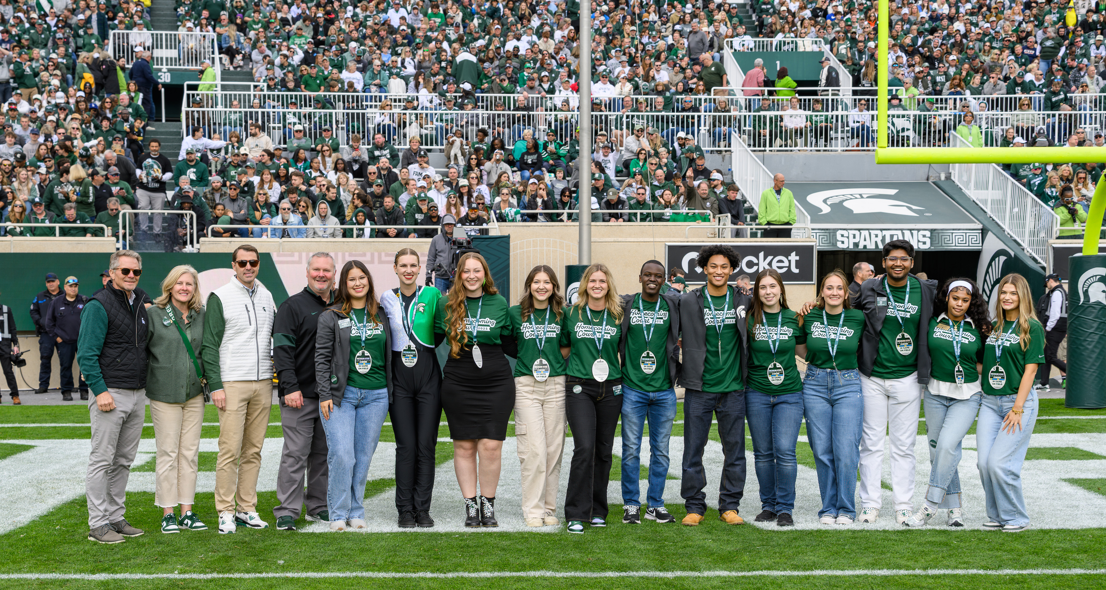 The 2025 MSU Homecoming Court stands on the football field at Spartan Stadium, smiling for a group photo in front of a packed crowd. Dressed in matching green Homecoming Court T-shirts and name badges, the students pose alongside university leaders and event organizers during a celebratory on-field recognition.