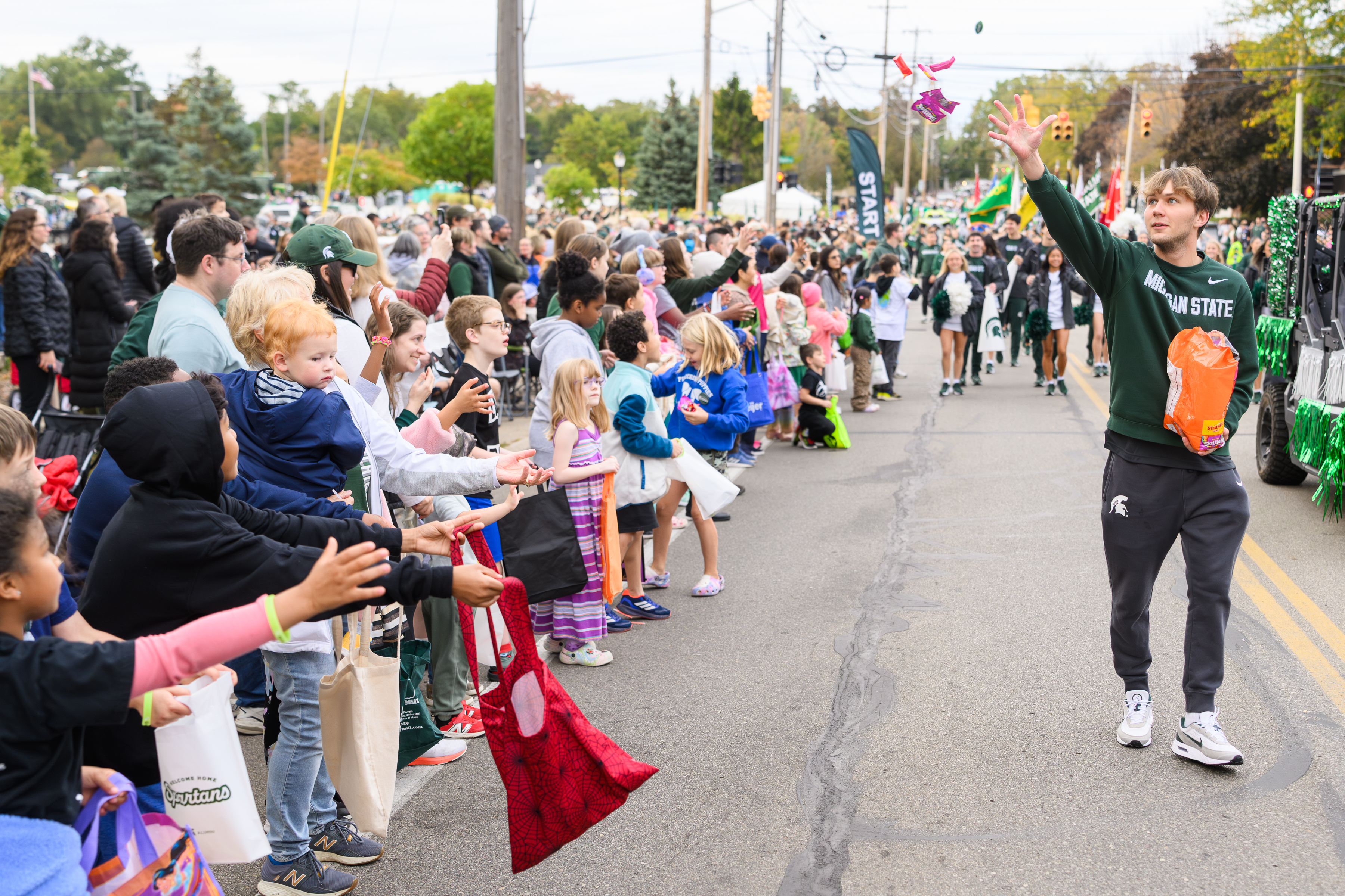 A Michigan State University student in a green Spartan sweatshirt tosses candy into a crowd of excited children and families lined up along the Homecoming parade route. Kids hold out tote bags and wear colorful outfits, eagerly reaching for treats. Behind them, more parade participants march down the street lined with green banners and festive decorations.