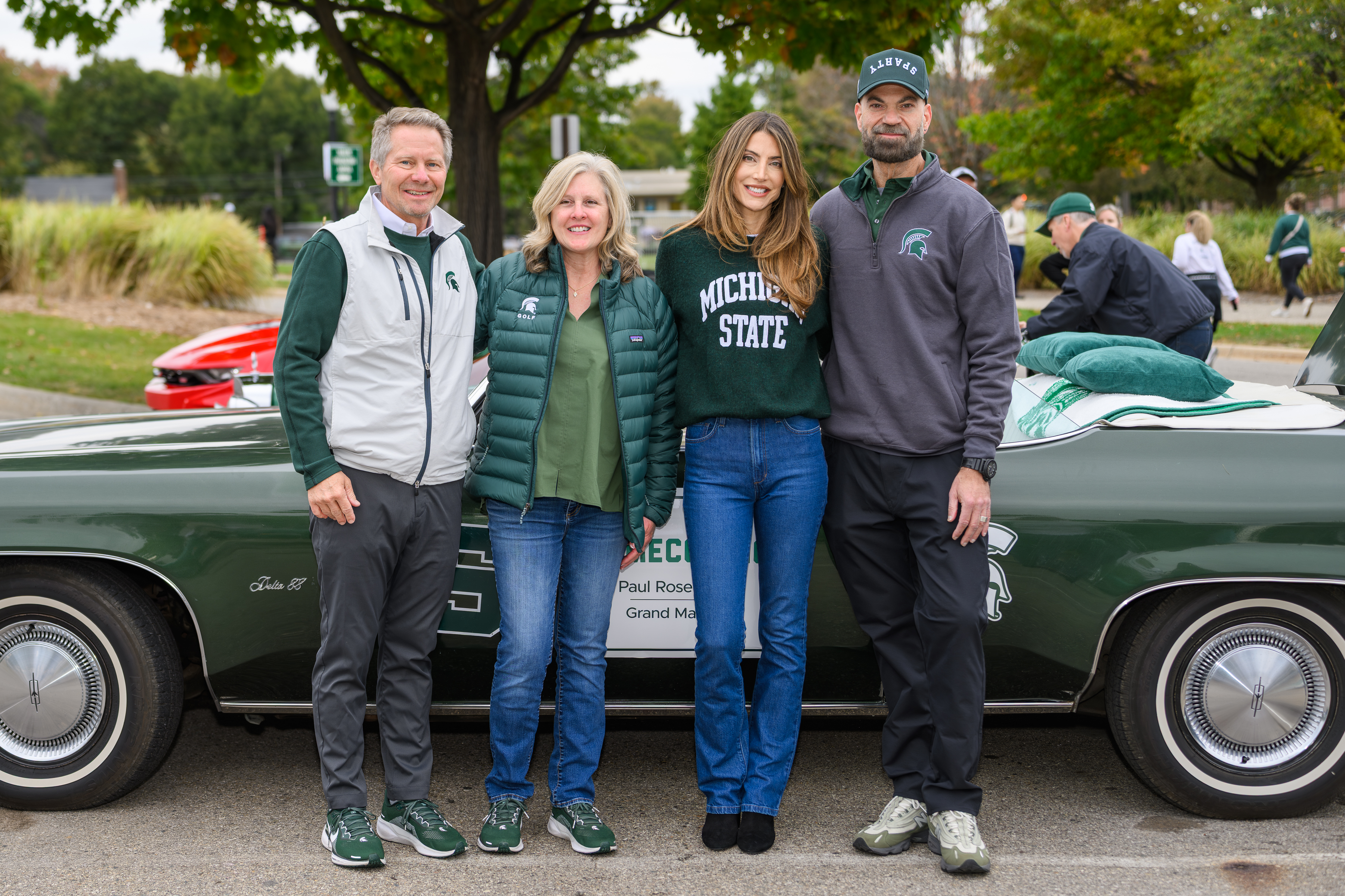 MSU President Kevin Guskiewicz, his wife Amy Guskiewicz, and Homecoming Grand Marshal Paul Rosenberg pose for a photo alongside a Michigan State alumna in front of a classic green convertible labeled “Paul Rosenberg – Grand Marshal.” All are dressed in MSU green and white apparel, smiling during Homecoming festivities with trees and parade activity in the background.