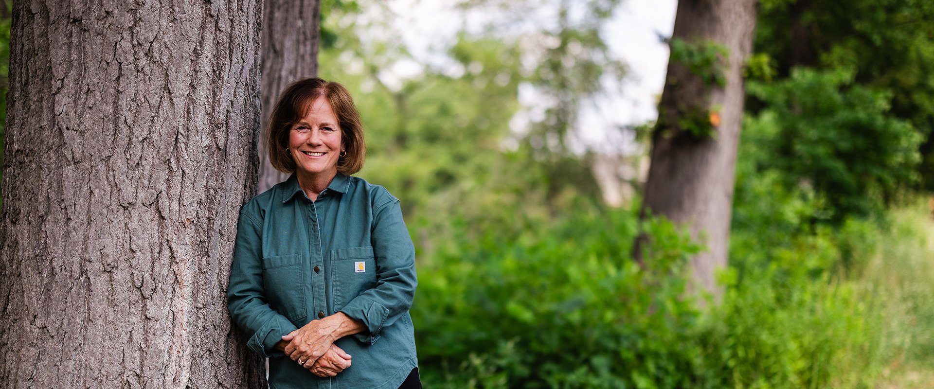 Linda Hubbard standing outdoors, leaning against a large tree trunk in a wooded area with green foliage and grass in the background.