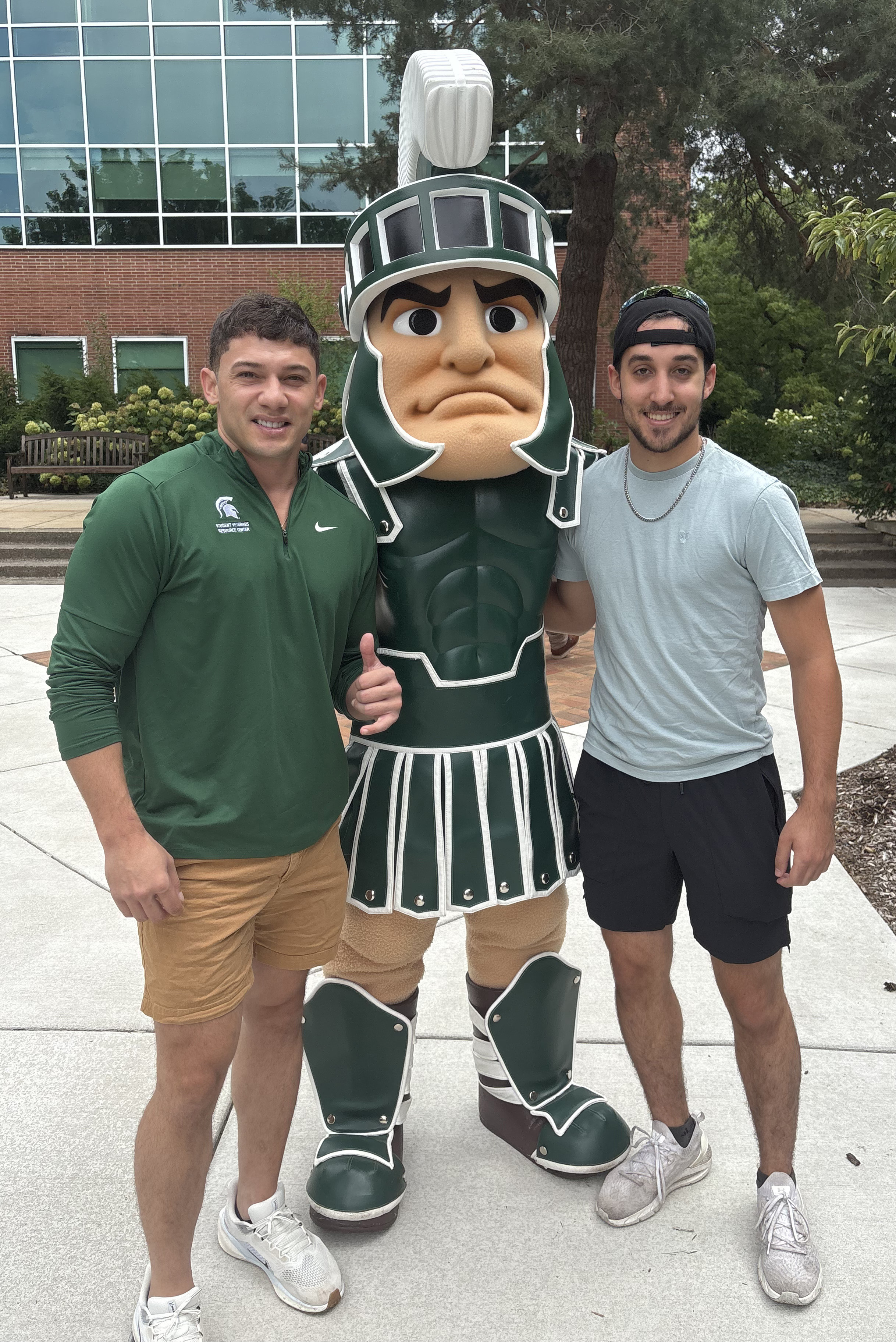 MSU student veterans Kyle Hines and Dom Pucillo pose for a photo with Sparty in front of a campus building.