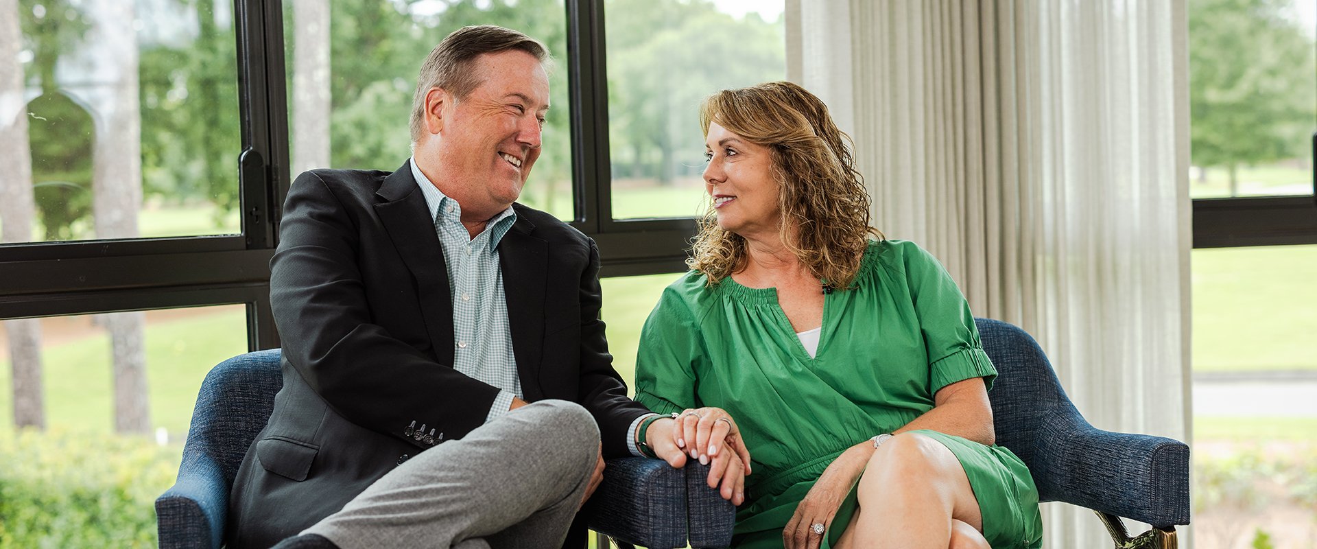 Mike and Mary Lamach seated indoors on blue chairs, holding hands near large windows with greenery outside.