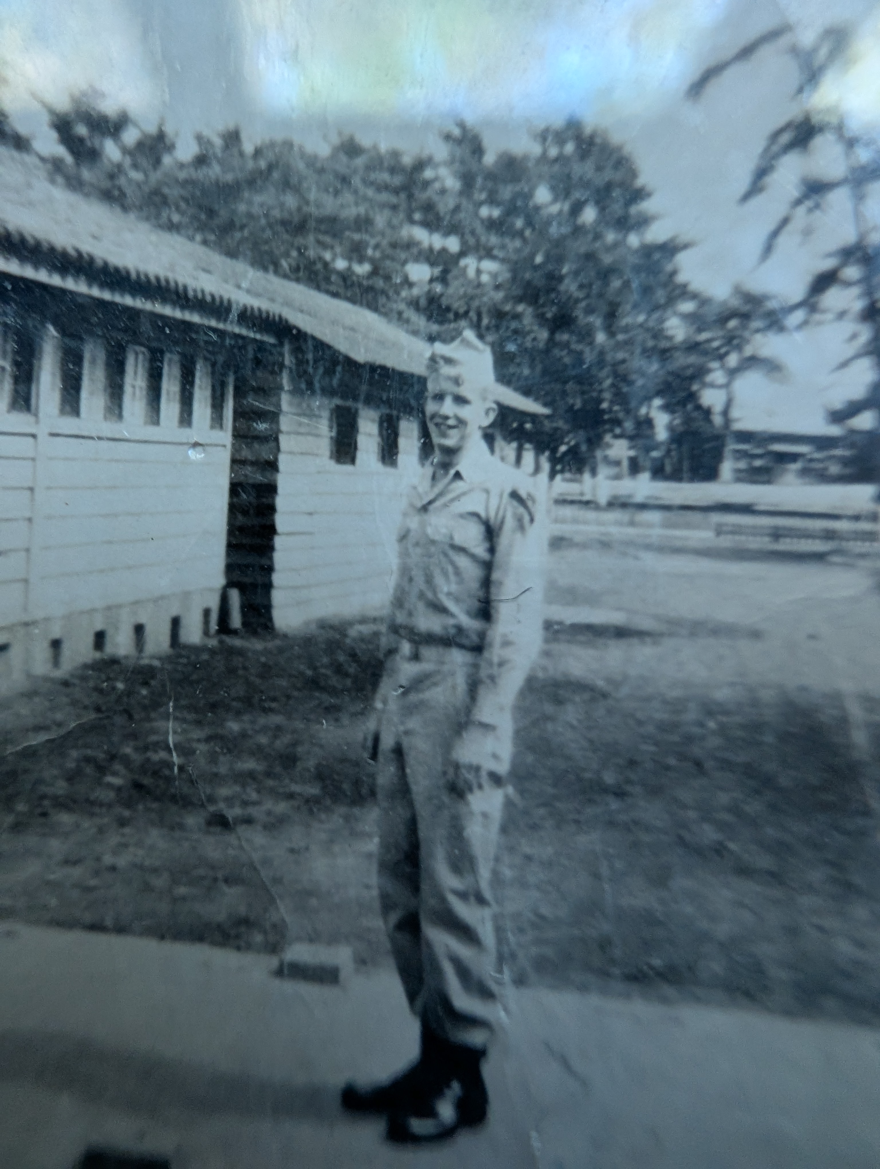 Black-and-white photo of a young man in uniform, smiling outside near wooden barracks with trees and buildings in the background.