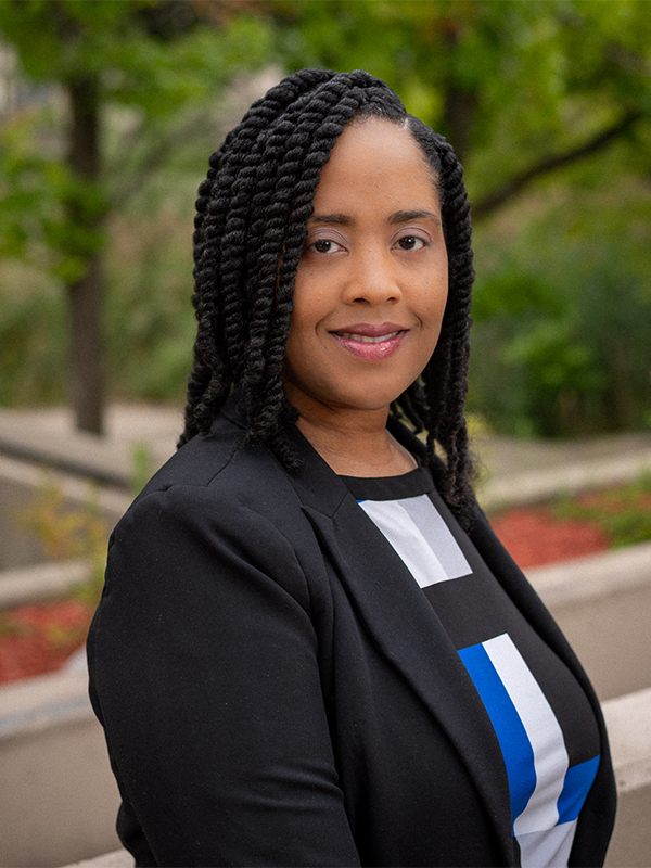 Essence, wearing a black and blue sweater and black blazer, smiling for a headshot. 