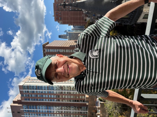 Jim Chetrick smiling on a city balcony wearing a Michigan State Spartans shirt and cap.