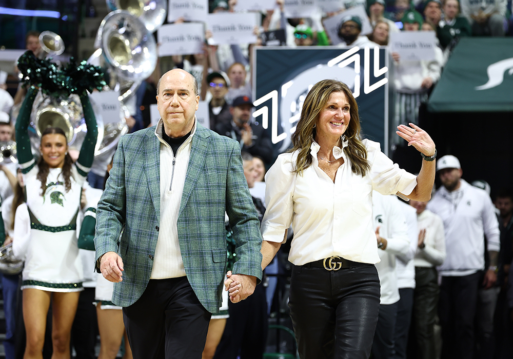 Dawn and Greg Williams holding hands while Dawn waves as they step onto the basketball court at Breslin Center