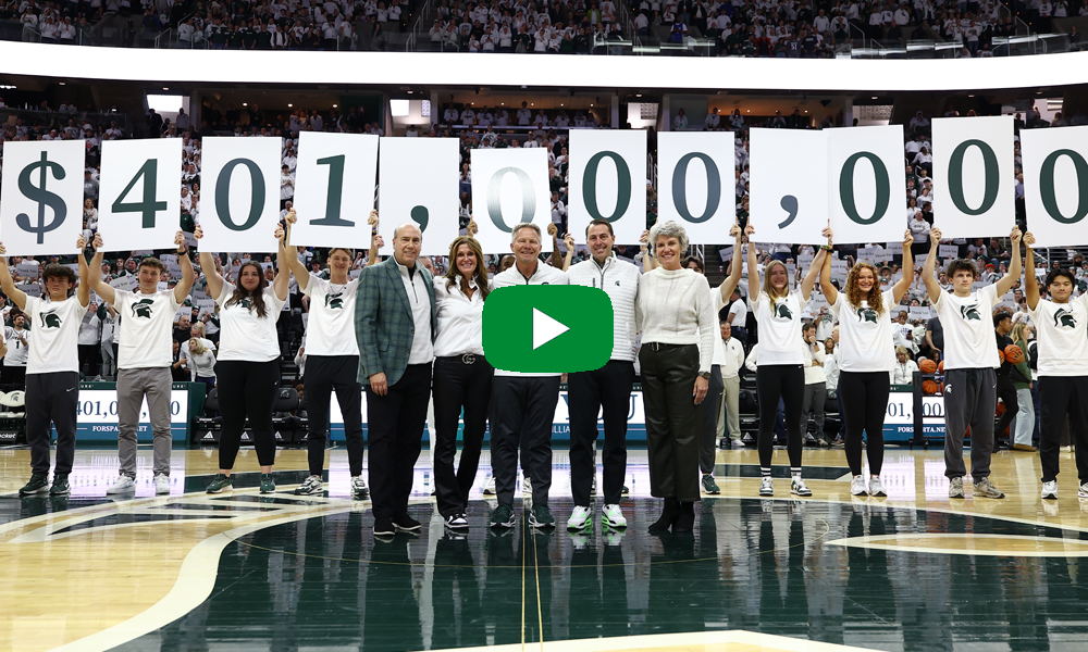 Greg and Dawn Williams and group on basketball court with students holding signs that display $401,000,000 during a large arena event.
