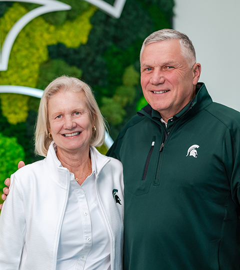 Jill and Jay Craig wearing MSU apparel stand together in front of a green wall with a large Spartan helmet logo.
