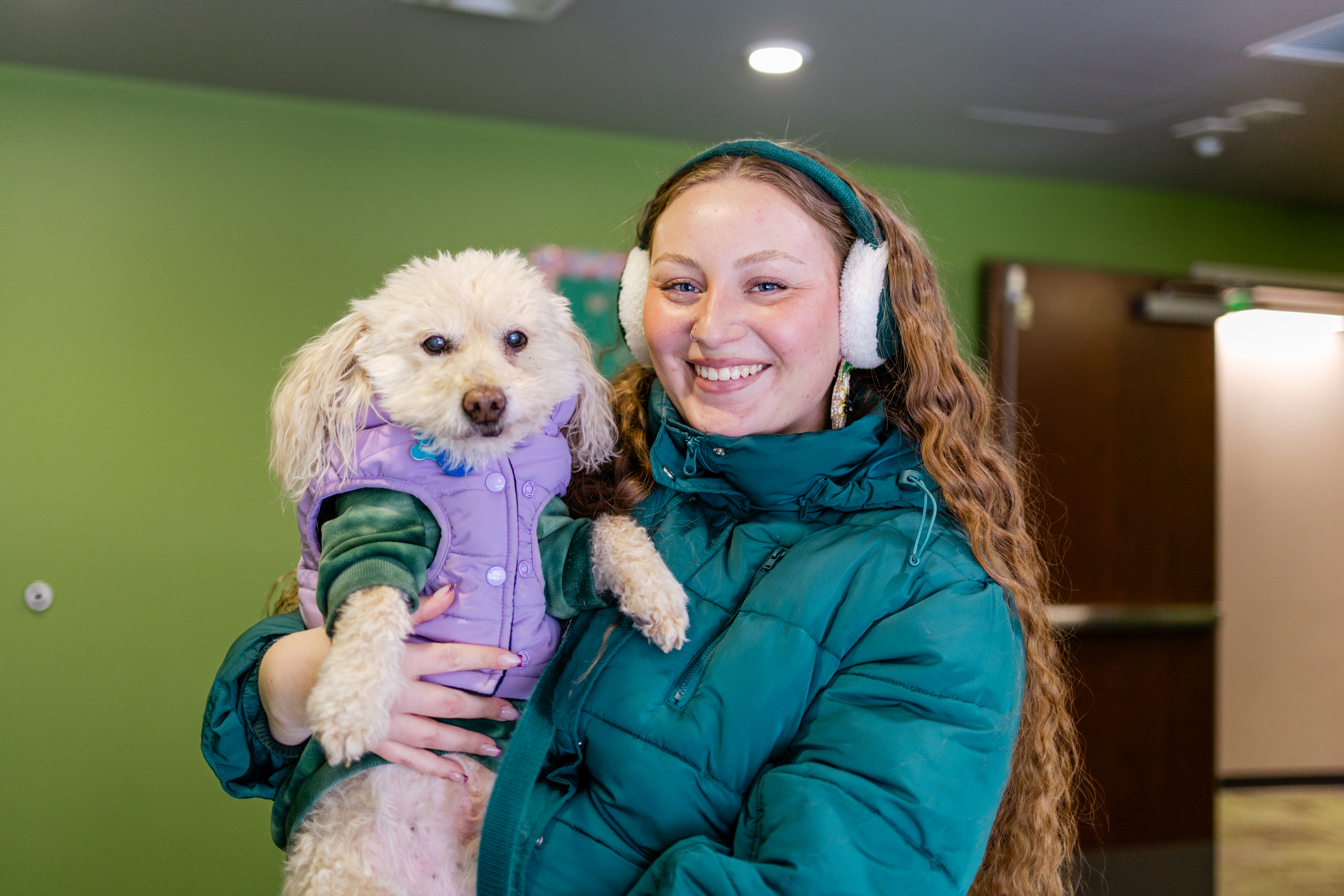Bundled up for winter, MSU student Kaylin Casper smiles at the camera while holding her dog in a campus hallway.