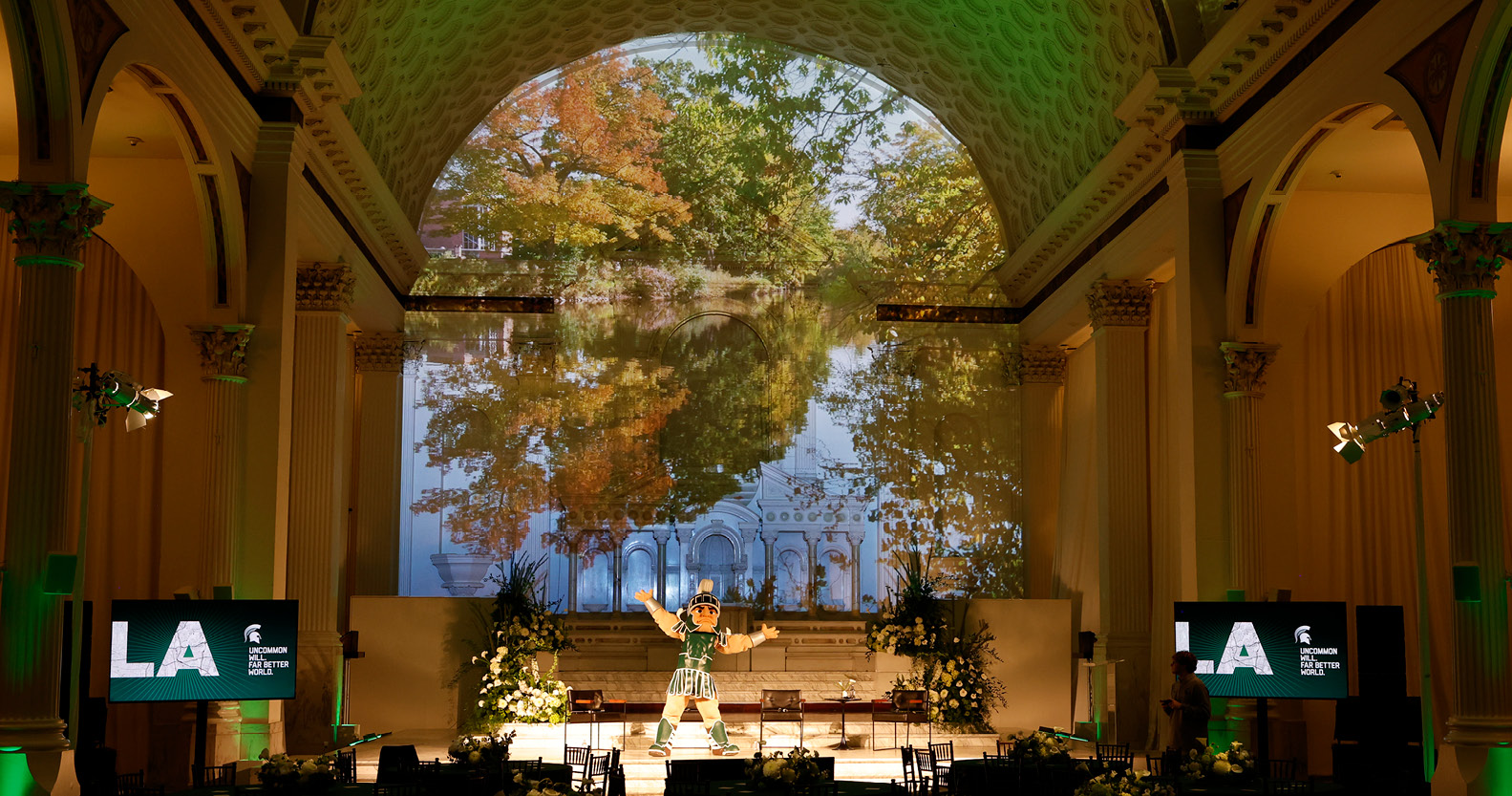 Interior of the event space featuring Sparty on stage in front of a projection of the Red Cedar River and fall trees on campus. 