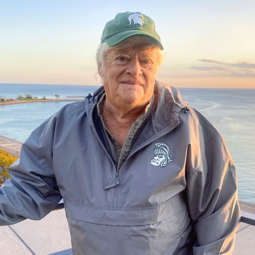Dave Spalding wearing MSU gear stands outdoors near water at sunset, with a shoreline and sky in the background.