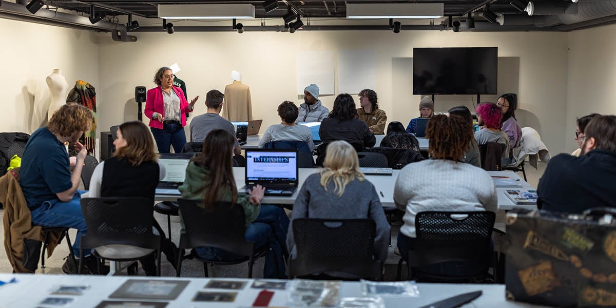 Professor Suzanne Fischer stands at the front of the Immersive Lab at the MSU Museum to teach her course on Museum Studies. A crowd of students sits in front of her at multiple tables.