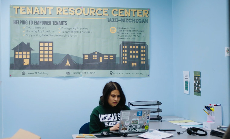Social Science Scholar Sumaiya Imad wearing an MSU sweatshirt sitting behind her laptop at a desk with a Tenant Resource Center banner in the background.