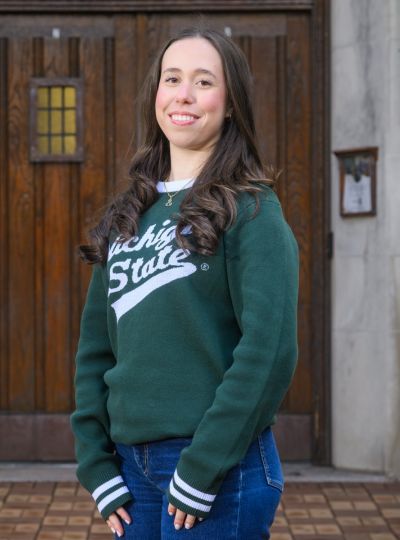 Headshot of a person in a green Michigan State sweatshirt and jeans, standing in front of wooden doors on a campus building.