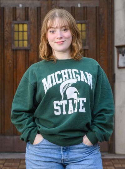 Person in green Michigan State sweatshirt and jeans standing in front of tall wooden doors with hands in pockets.