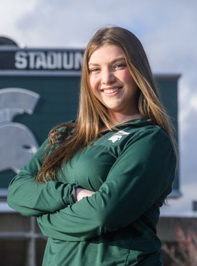 Person wearing Michigan State apparel stands with arms crossed in front of the Spartan Stadium scoreboard.