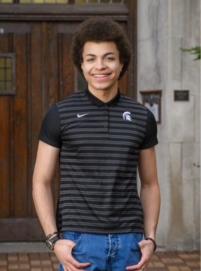 Person wearing a black striped Michigan State polo stands in front of large wooden doors on a stone building walkway.