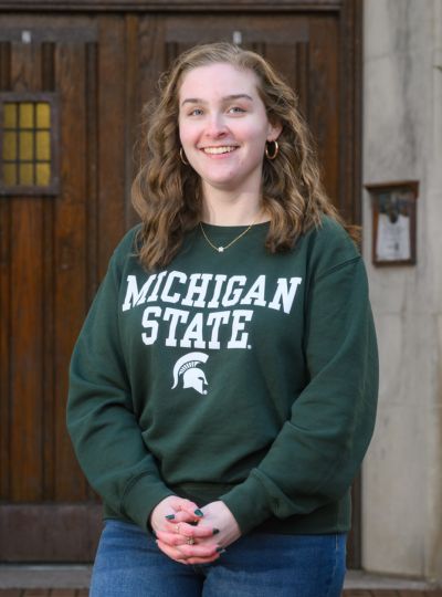 Person wearing a green Michigan State sweatshirt stands with hands clasped in front of large wooden doors on a stone building.