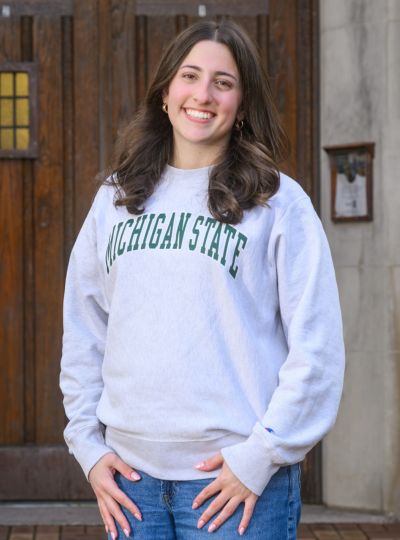 Person wearing a light gray Michigan State sweatshirt stands with hands at sides in front of large wooden doors on a stone building.