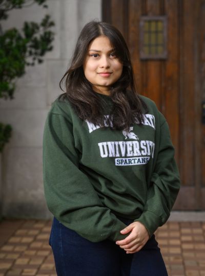 Person wearing a green Michigan State University Spartans sweatshirt stands with hands clasped in front of large wooden doors on a stone building.