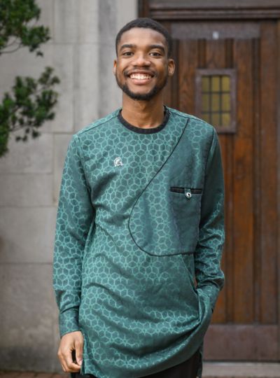 Person wearing a green patterned Michigan State long-sleeve top stands with one hand in a pocket in front of large wooden doors on a stone building.