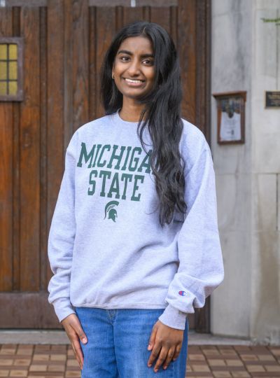 Person wearing a light gray Michigan State sweatshirt stands with hands at sides in front of large wooden doors on a stone building.