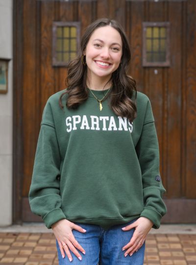 Person wearing a green Michigan State Spartans sweatshirt stands with hands at sides in front of large wooden doors on a stone building.