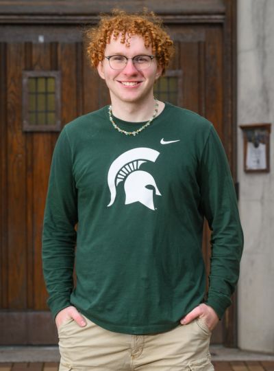 Person wearing a green Michigan State long-sleeve shirt with Spartan helmet logo stands with hands in pockets in front of large wooden doors on a stone building.