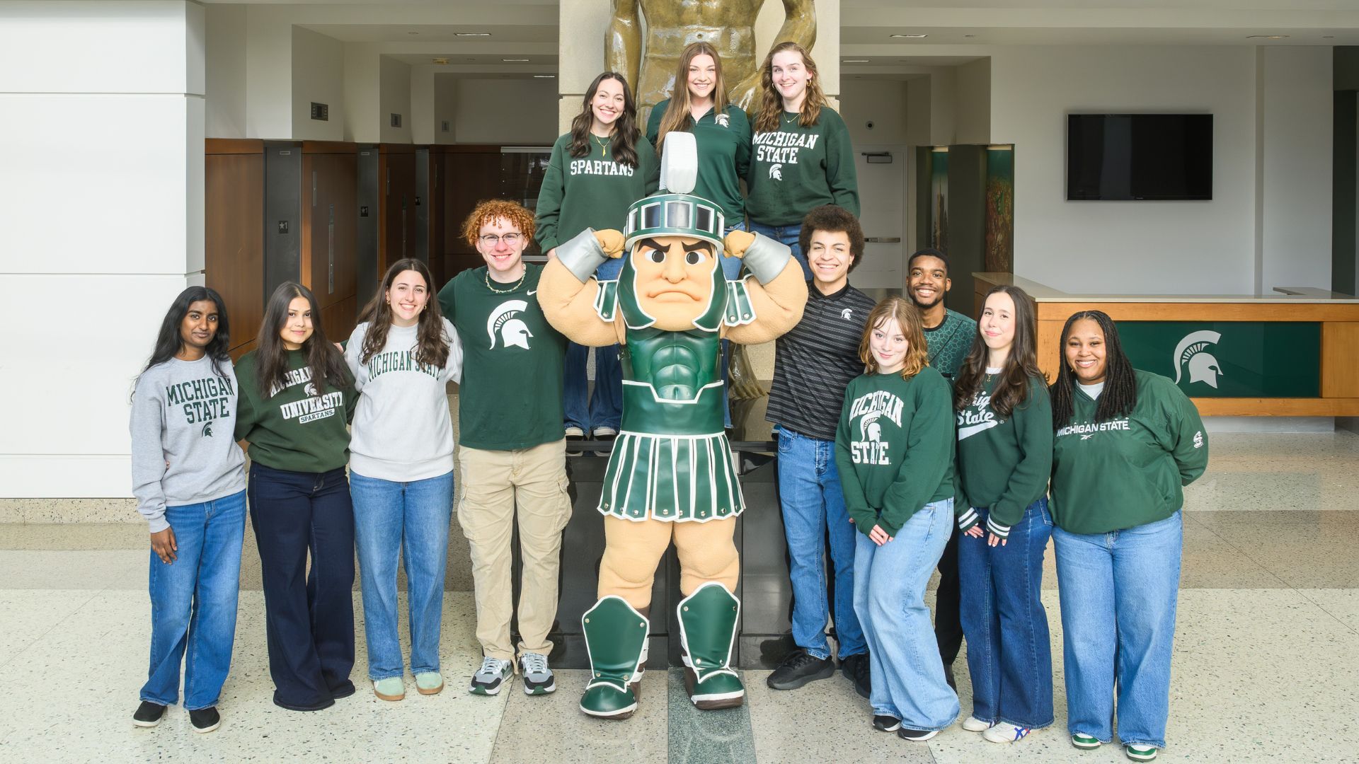 Group of Michigan State students in green and white stand around the Sparty mascot, posing together inside a campus building lobby.
