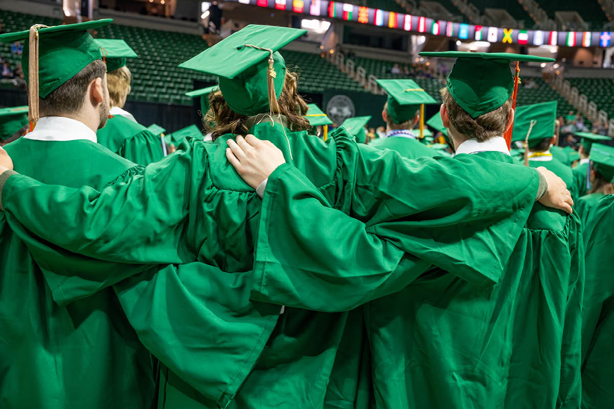 Michigan State graduates in green caps and gowns stand with their arms around one another during commencement.