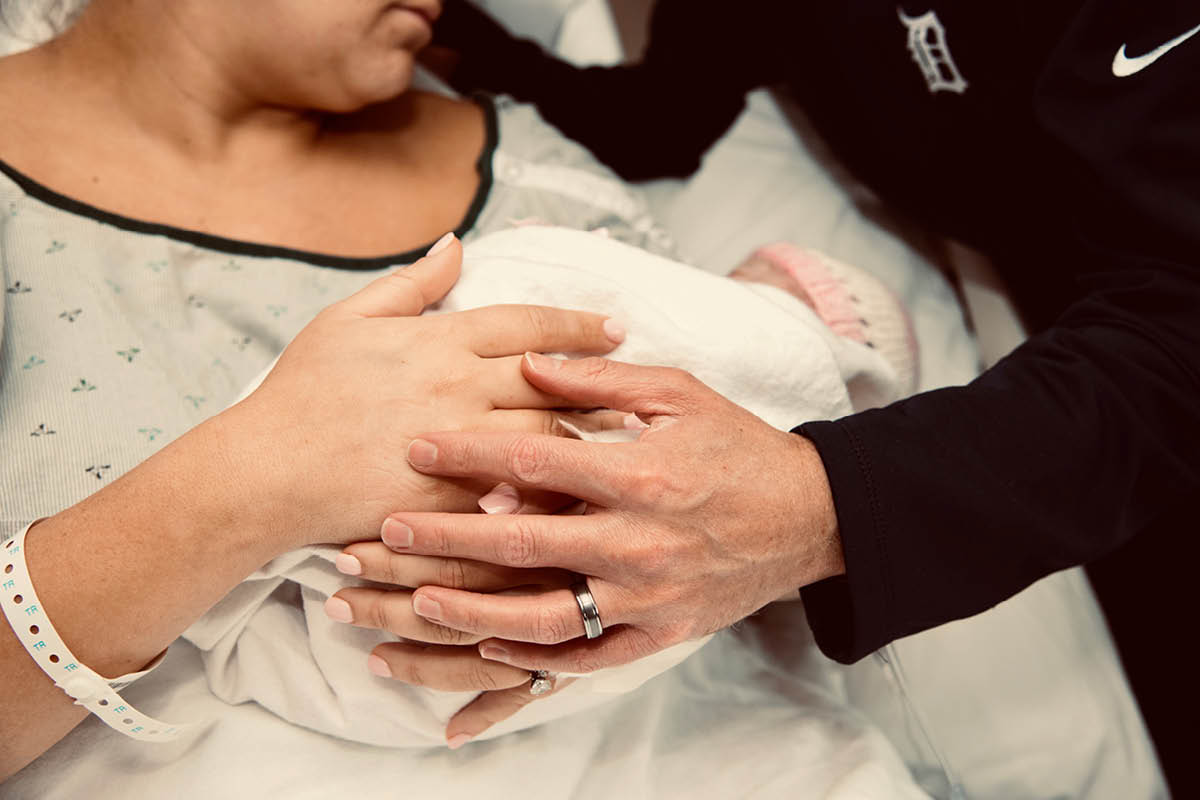 A mother in a hospital gown holds a newborn baby wrapped in blankets, with the father’s hand resting on top of hers in support.