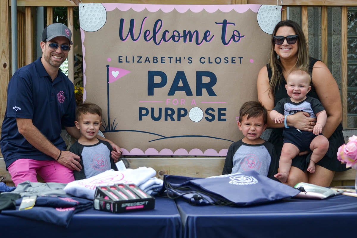 Jeff and Elizabeth Bearance sit at a community golf event with their three sons in front of a sign that reads: