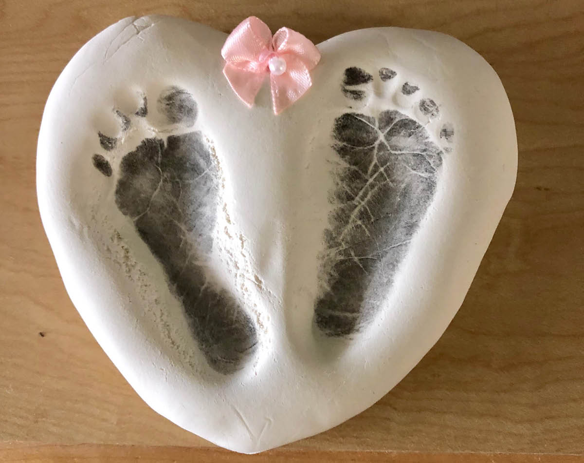 A white heart-shaped plaster cast of two small baby footprints with a small pink satin ribbon bow at the top.
