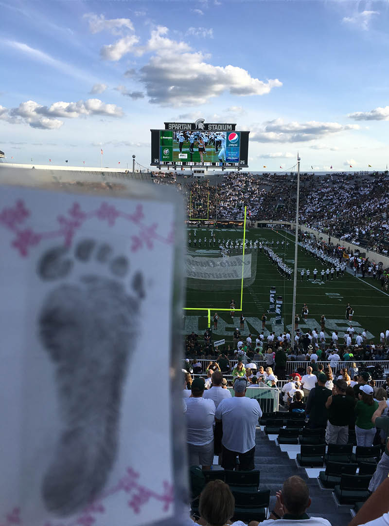 A card featuring a baby footprint is held up in the foreground with a crowded Spartan Stadium in the background while a football games is played on the field.