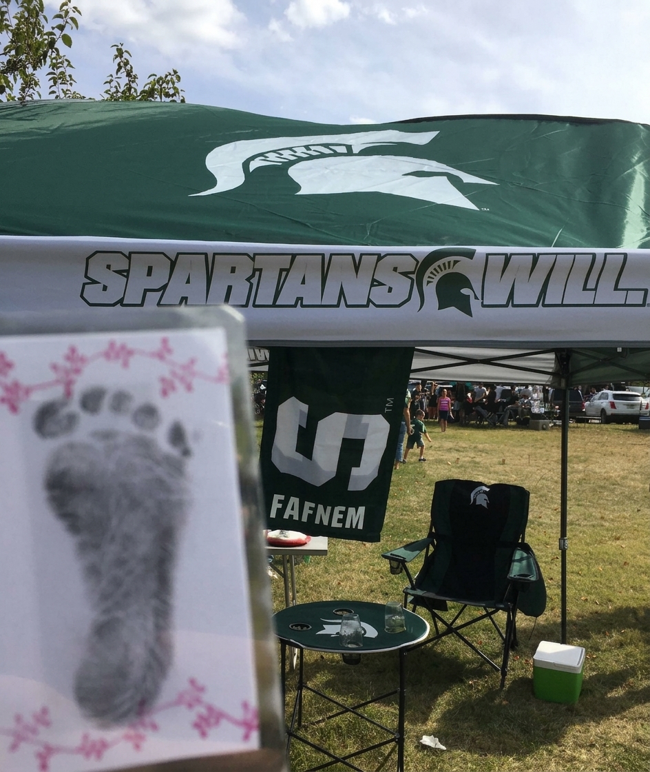 A card featuring baby footprints is held up in the foreground with an outdoor tailgate scene with a Michigan State Spartans canopy tent in the background.