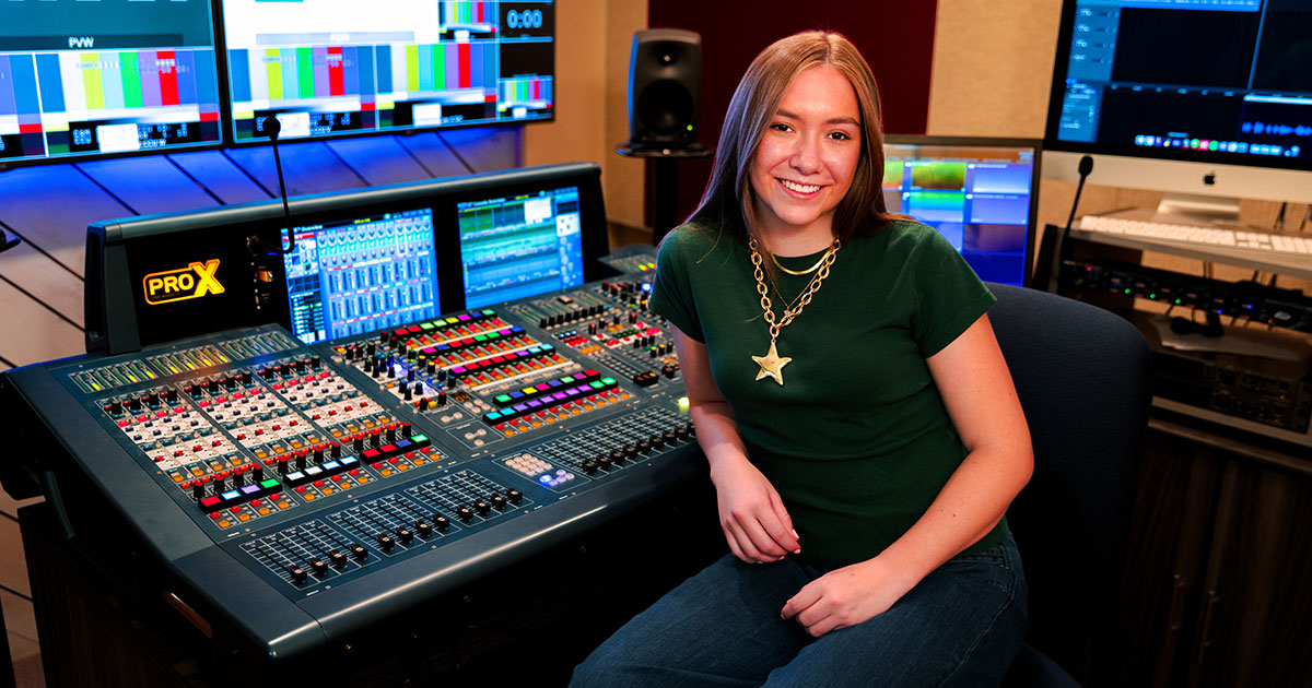 Ava Crozier sits beside a professional audio mixing console in a multimedia production studio surrounded by monitors and equipment.