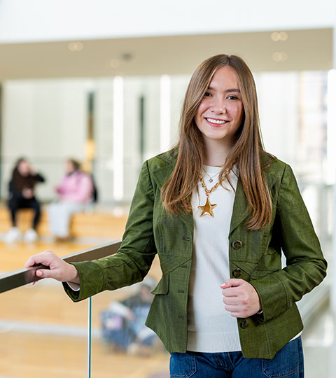 Ava Crozier in a green blazer poses indoors, leaning on a railing in a bright, modern campus building.