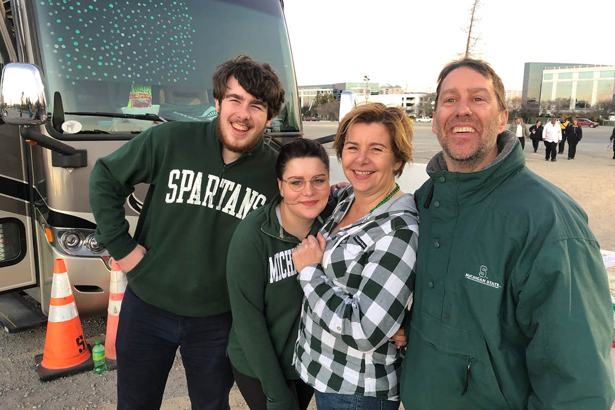Constance Laflamme stands in an outdoor parking lot smiling with her husband and two adult children, all wearing Michigan State apparel.
