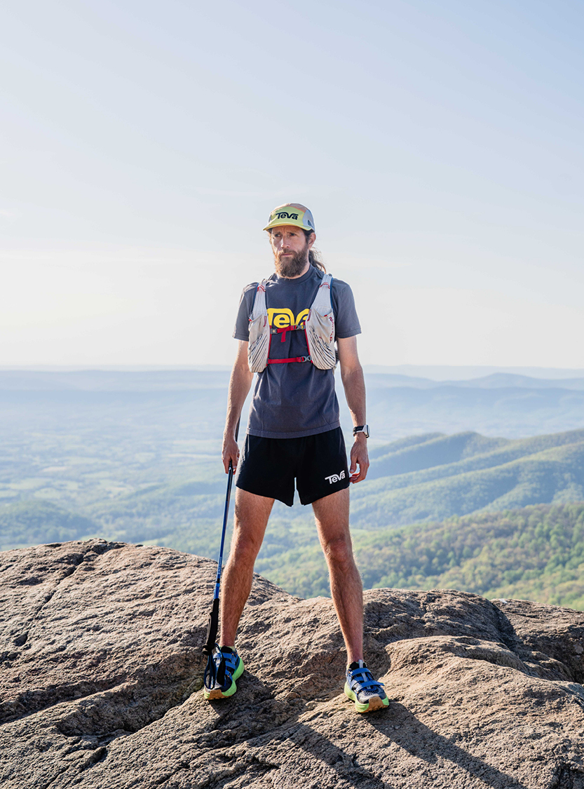 Mike Wardian on a mountaintop wearing a hat, vest and carrying walking sticks. 