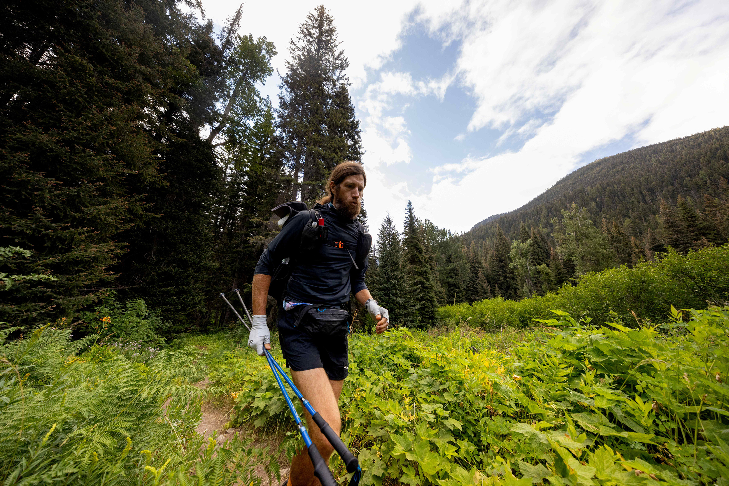 Ultrarunner Mike Wardian walking through a clearing in the woods with pine trees and a blue sky in the background.