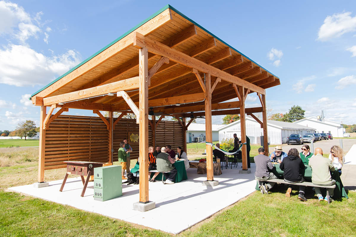 An outdoor gathering featuring a large, wooden beam pavilion with a slanted roof over a concrete slab...