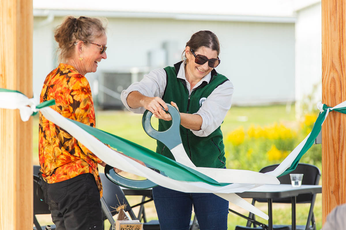Dr. Meghan Milbrath in a green MSU vest uses giant ceremonial scissors to cut a green and white ribbon while donor Charlotte Hubbard, wearing an orange floral shirt and sunglasses, smiles alongside her under a wooden shelter.