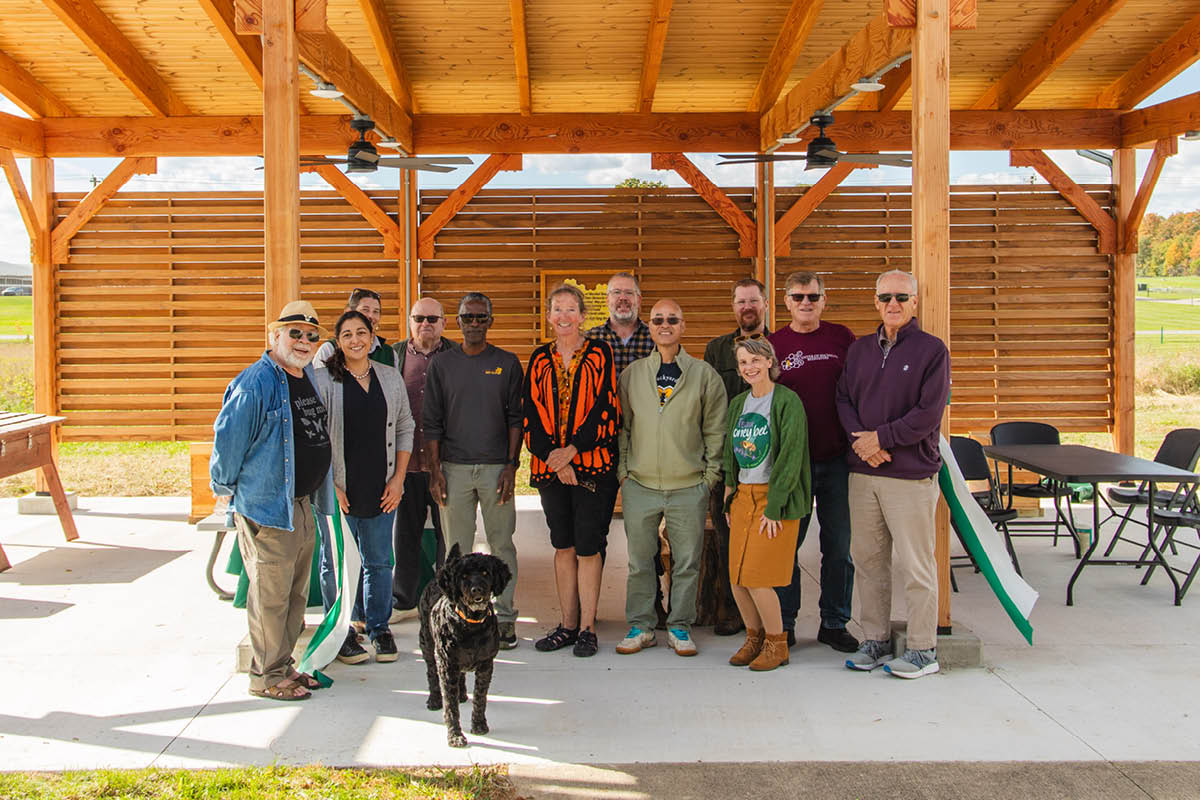 A group pose of attendees, including Charlotte Hubbard and a black dog, standing together under the wooden teaching pavilion at the dedication event.