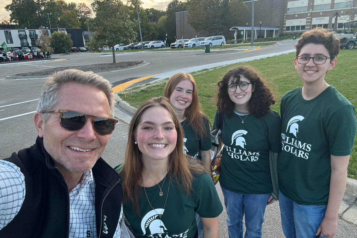 MSU President Kevin Guskiewicz (left) takes an outdoor selfie with four smiling Williams Scholarship recipients dressed in matching green MSU Williams Scholars t-shirts.