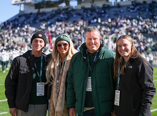 Four members of the Zeigler family in green and black Spartan apparel pose together on the field at a crowded Spartan stadium.
