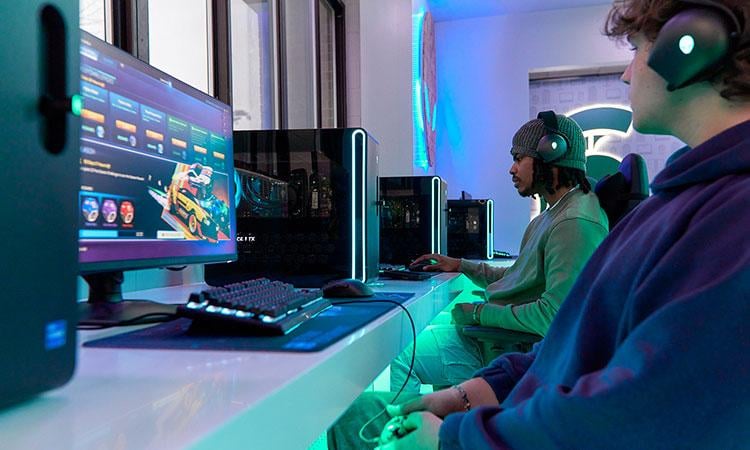 Students wearing earphones and holding game controllers play PC-based games on a row of desktop computers.