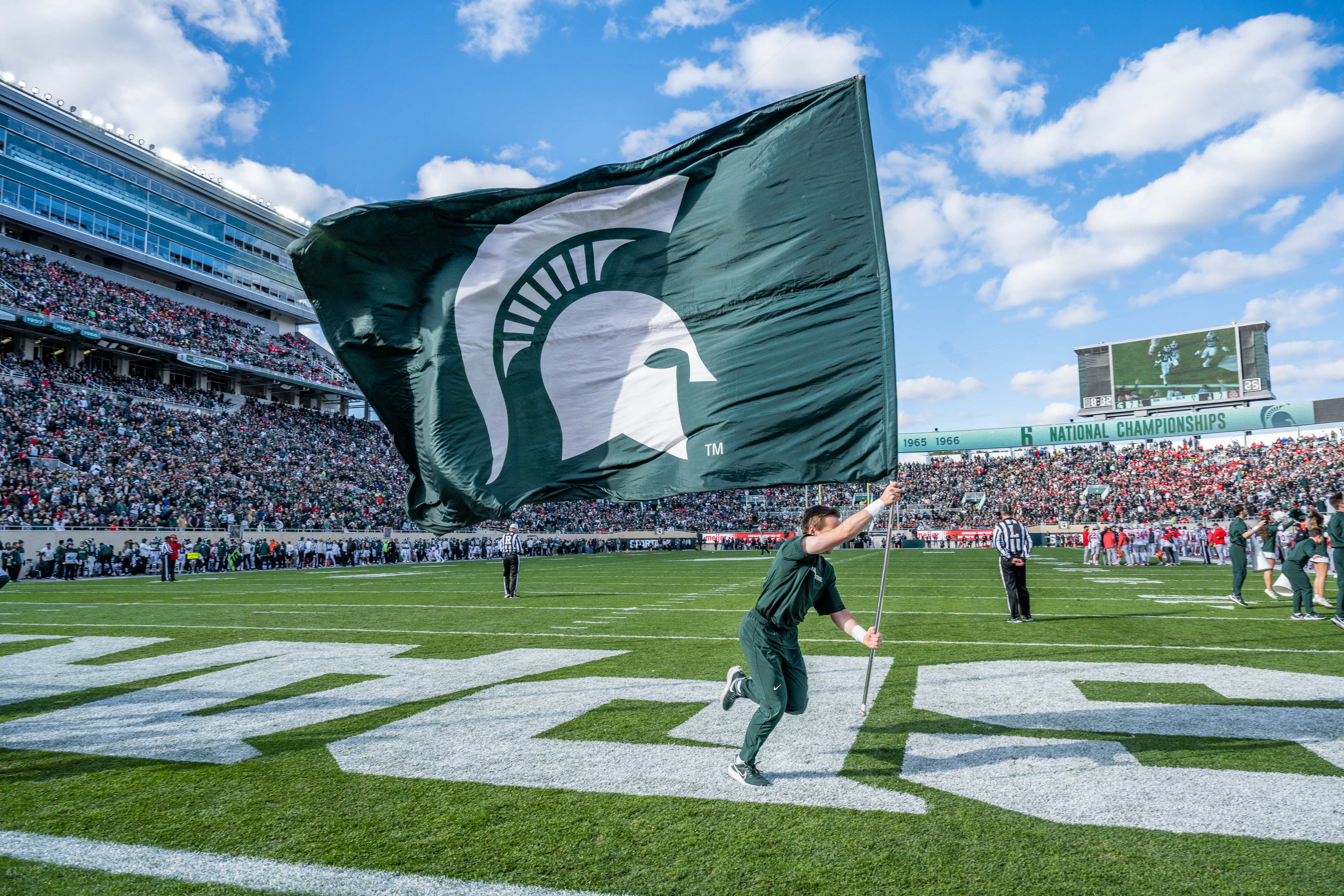 This image captures the excitement of a Michigan State University football game. A student, dressed in green, is seen running across the field while holding a large Spartans flag, which prominently displays the iconic white Spartan helmet logo on a green background. The crowd in the stadium is packed, with fans filling the stands, creating a vibrant atmosphere. The sky is bright with scattered clouds, and a large scoreboard is visible in the background, showing the game in progress. The energy of the scene highlights the spirited support for the Spartans during the game.
