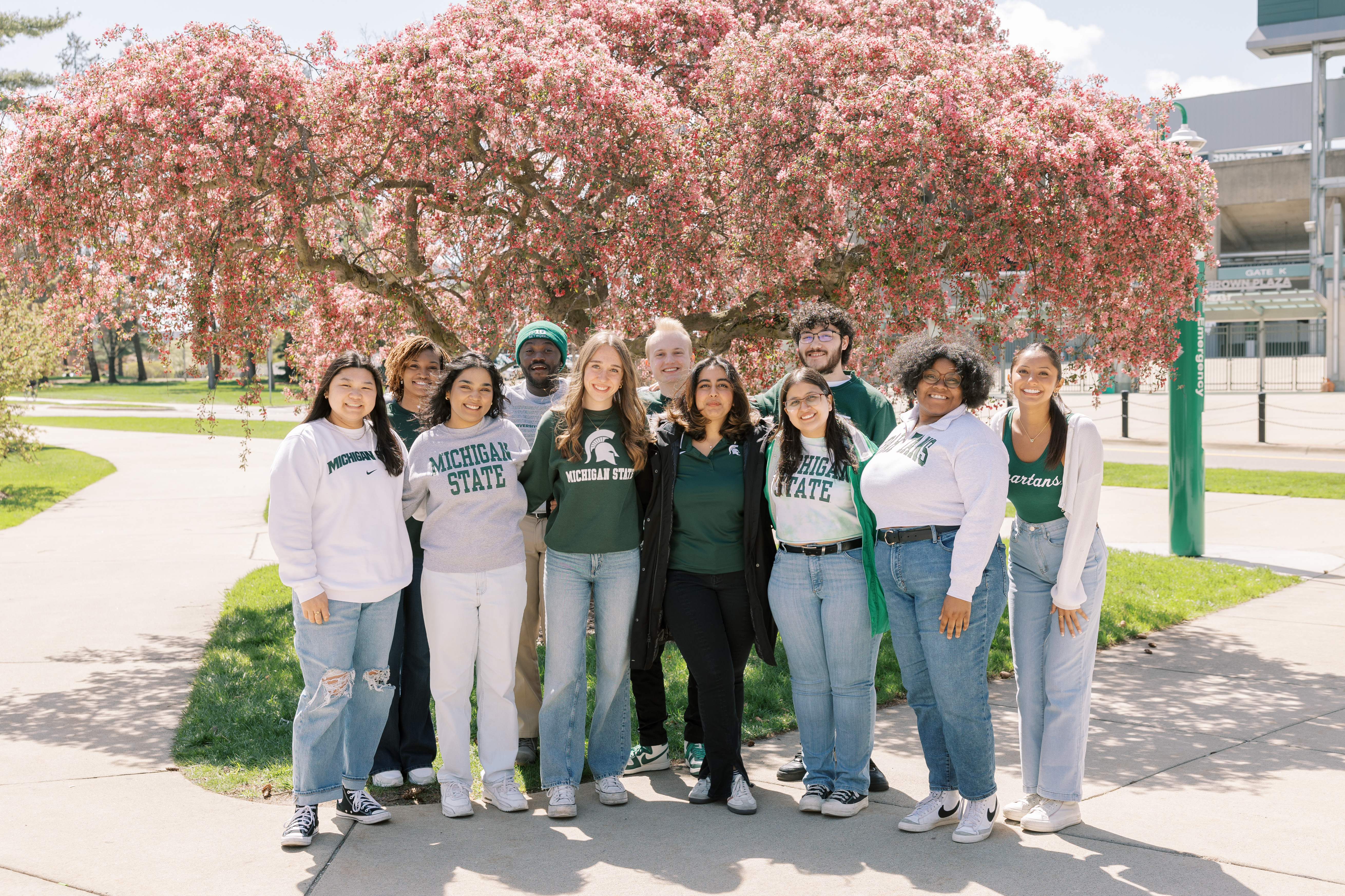This image shows the Michigan State University Homecoming Court standing together in front of a blooming tree with vibrant pink flowers. The group consists of individuals all dressed in Michigan State attire, including sweatshirts and t-shirts in shades of green, gray and white. They are standing close together, smiling and enjoying the sunny day on campus. The background features a well-manicured lawn and part of the stadium, creating a picturesque and welcoming scene that highlights the spirit and camaraderie of the Homecoming Court.